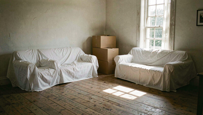An empty farmhouse living room with two sofas under white dust sheets, plain cardboard boxes stacked in the corner beside a tall window, afternoon sun across a worn wooden floor